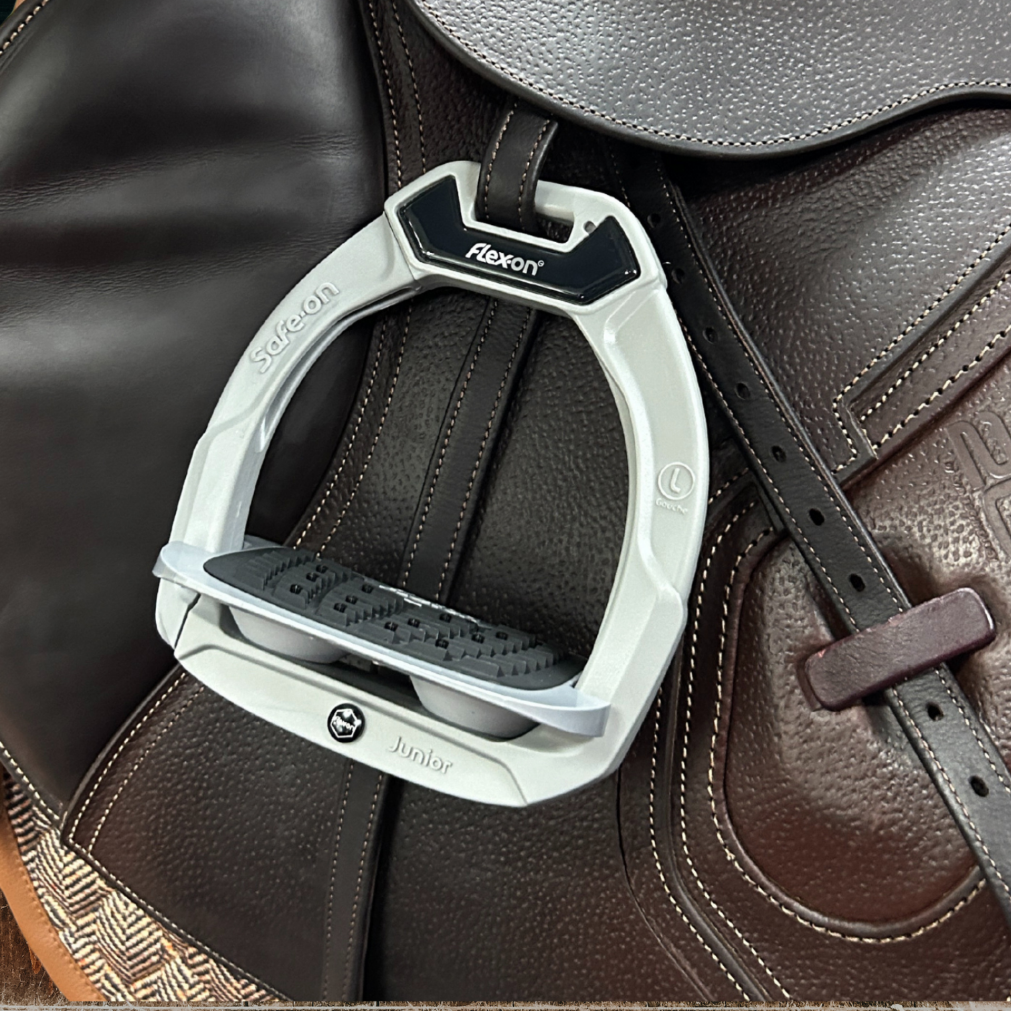 A Flex-on Safe-On Junior Stirrup with an inclined grip, featuring a grey color and organically sourced polyamide outer with a black leather riding boot and saddle in the background.
