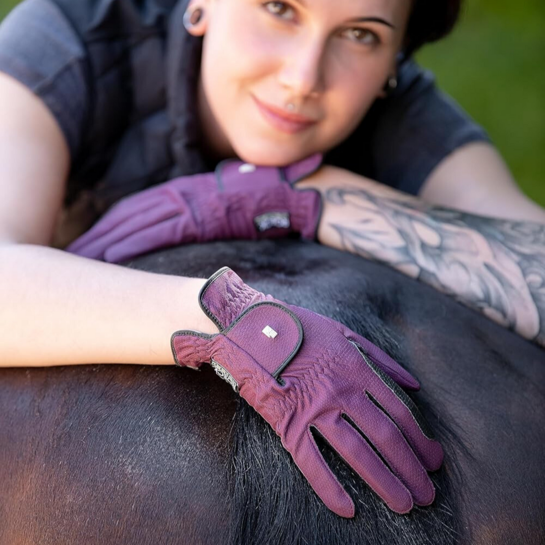A woman wearing purple Roeckl Lona gloves, designed for equestrian riding, resting her elbow on a horse with visible touchscreen compatibility on the gloves.