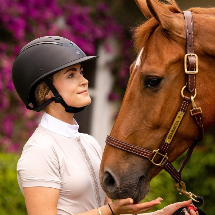 Lady wearing a EQx by Charles Owen Kylo Black Matte with Black Gloss Frame Riding Helmet. She is holding a chestnut horse wearting a brown leather halter.