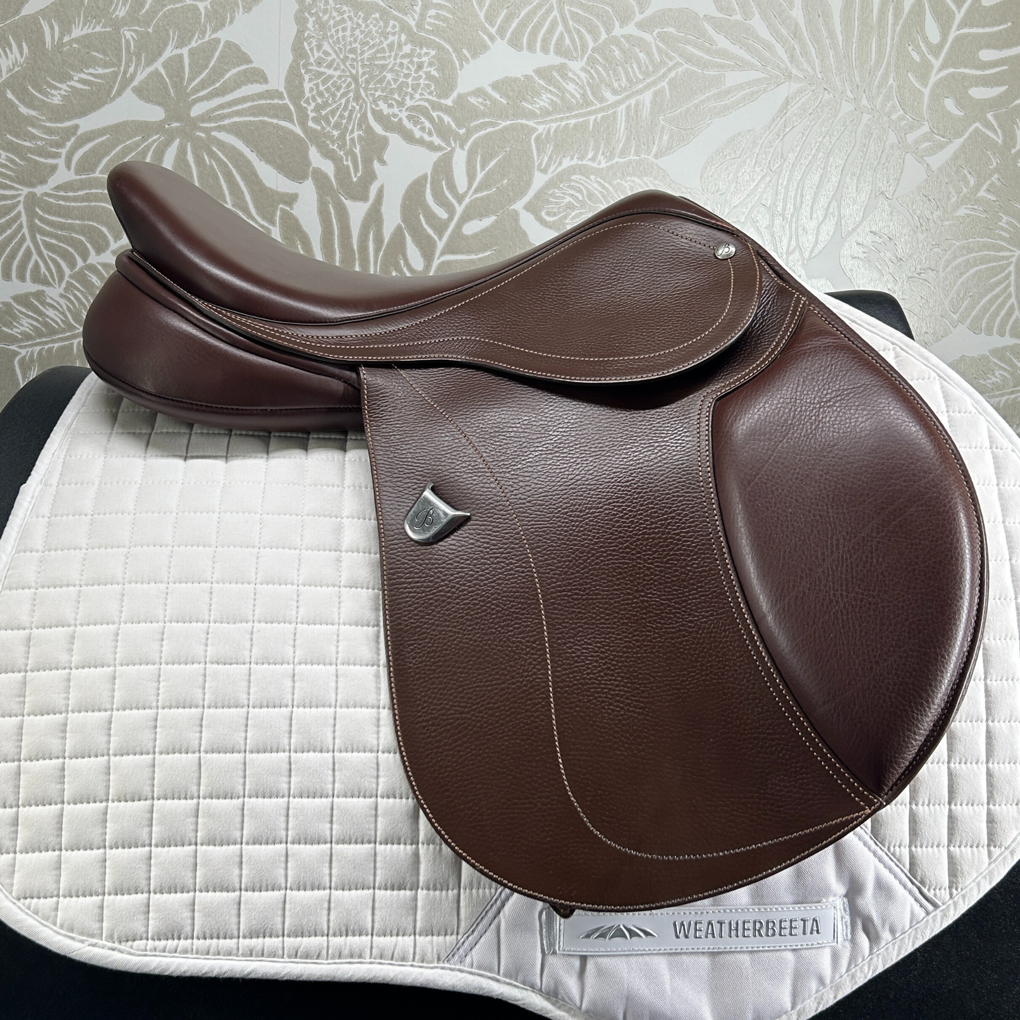 Brown used 17" Bates Hunter Jumper saddle on a white quilted pad with against a floral-patterned wall.