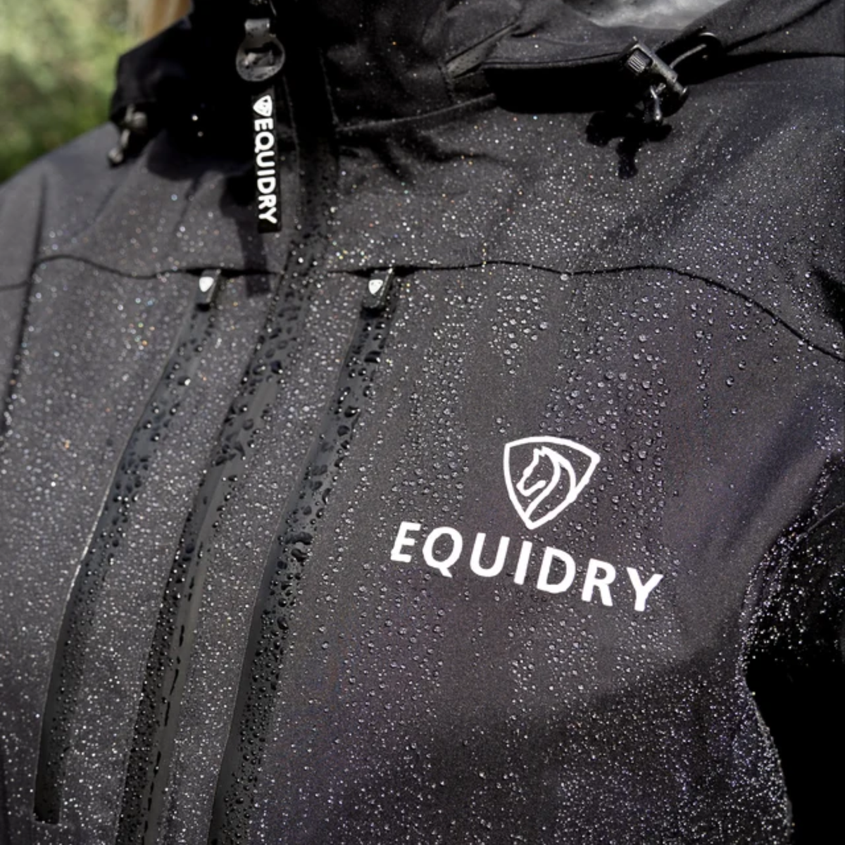 Close-up of a black EQUIDRY waterproof Varsha coat covered in water droplets, showing the white EQUIDRY logo on the chest and a black hood drawcord with white branding.