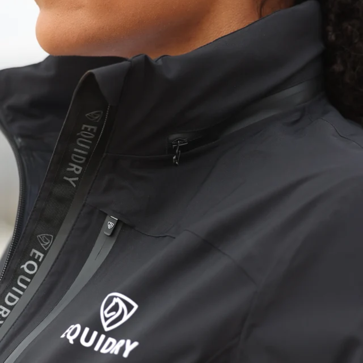 Close-up of a person wearing a black EQUIDRY Varsha coat, showing the collar, zipper, and white Equidry logo on the front.