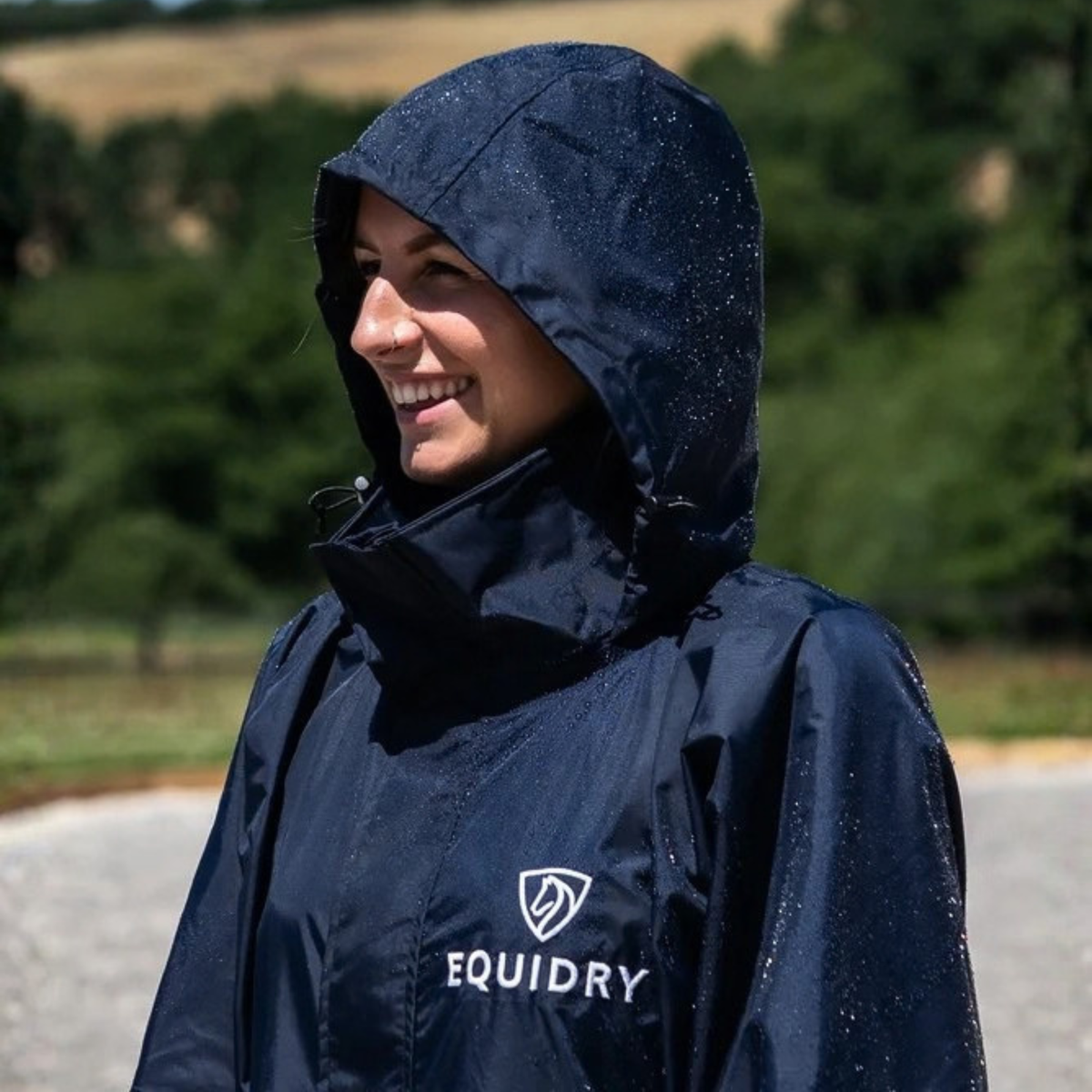 A a close up of awoman wearing a navy EquiDry Evolution Mesh Lined Coat with a high hood and collar, standing outdoors on a gravel path with trees in the background.