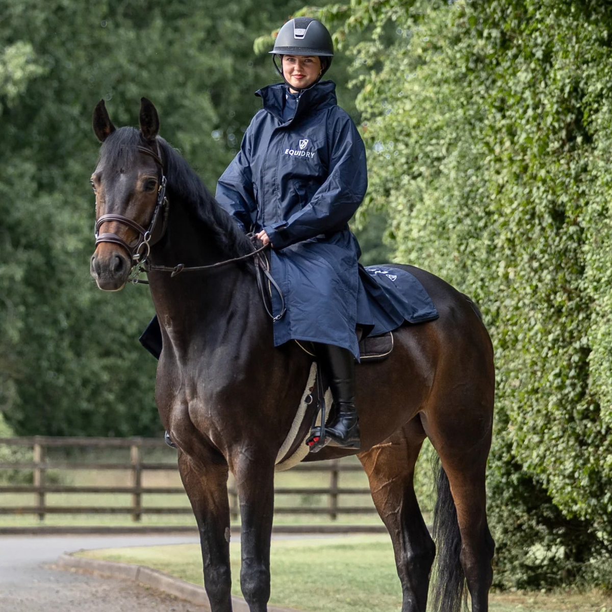 A rider wearing a navy blue EquiDry Evolution Mesh Lined Coatt and helmet rides a dark brown horse outdoors on a path with green trees in the background. The raincoat has a white "EquiDry" logo on the chest.