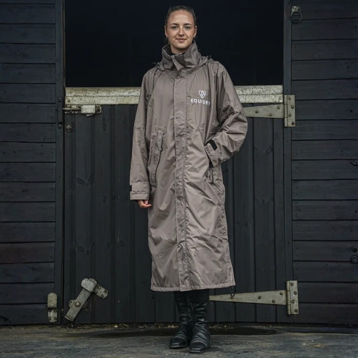 A woman stands in front of a wooden stall door wearing a long taupe EquiDry Evolution Mesh Lined waterproof coat with a white &