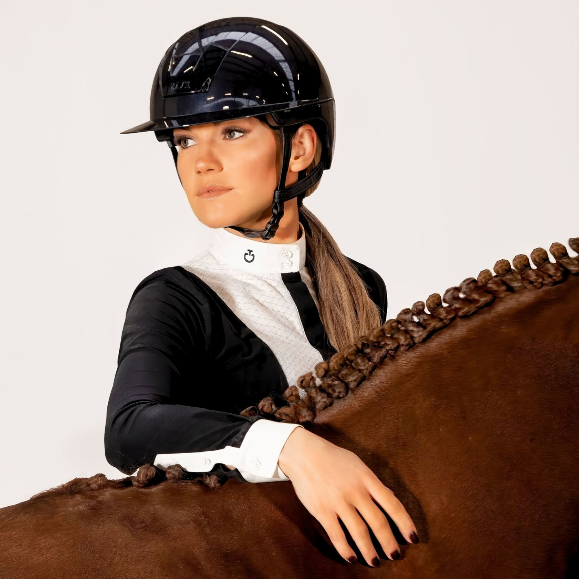 A woman in equestrian attire wearing a glossy black KASK Kooki Lady riding helmet and a black-and-white show shirt, posed with a brown horse against a plain light background.