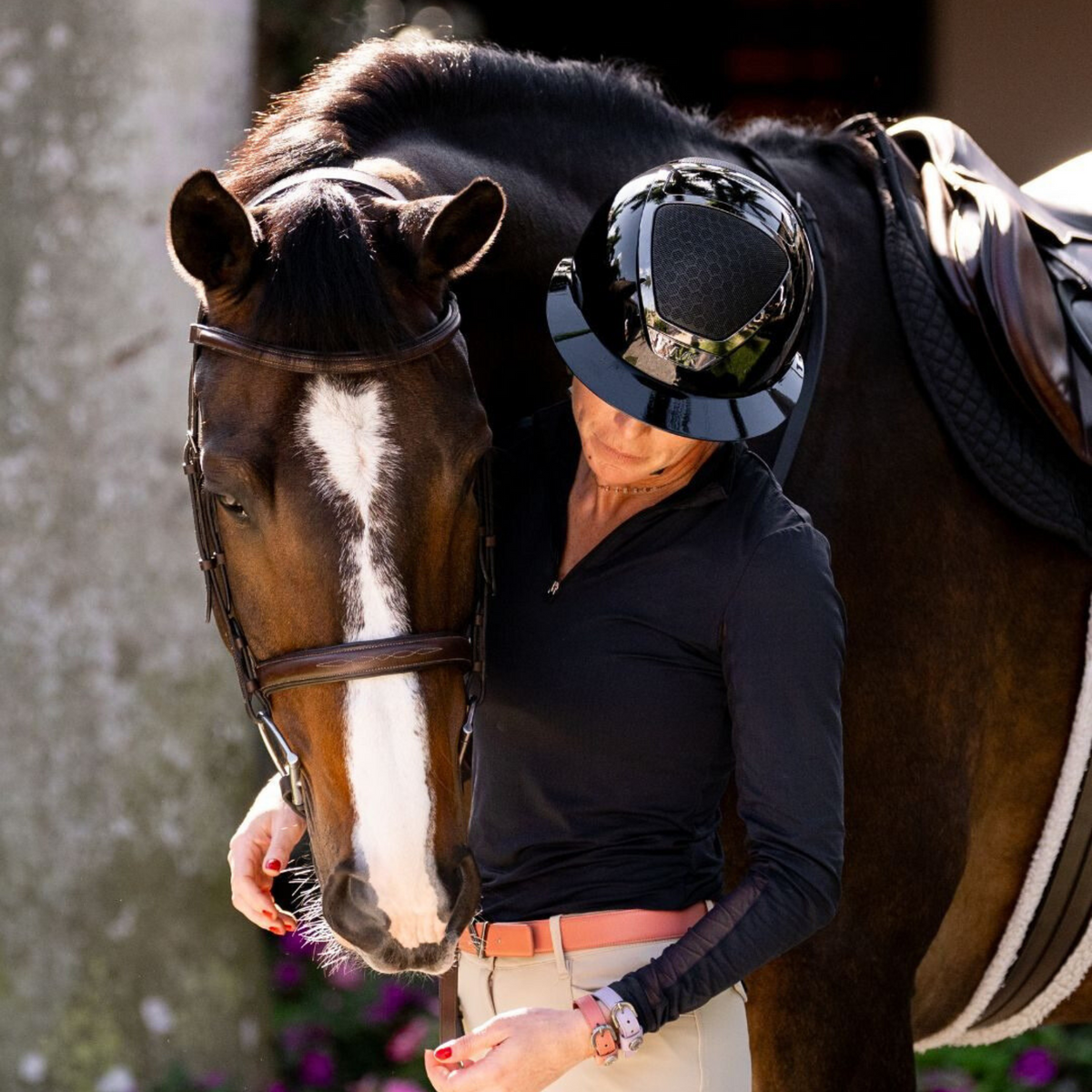Woman in equestrian attire wearing a black KASK Star Lady equestrian helmet standing next to a horse with a white blaze.