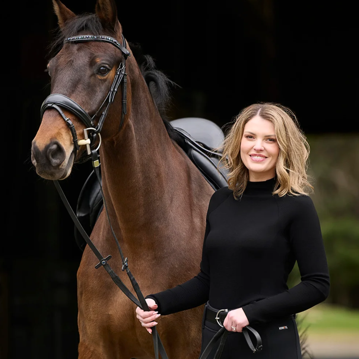 A woman wearing Kerrits black long-sleeve Align Textured Mock Neck top stands beside a brown horse wearing a black saddle and bridle, holding reins in outdoor setting.