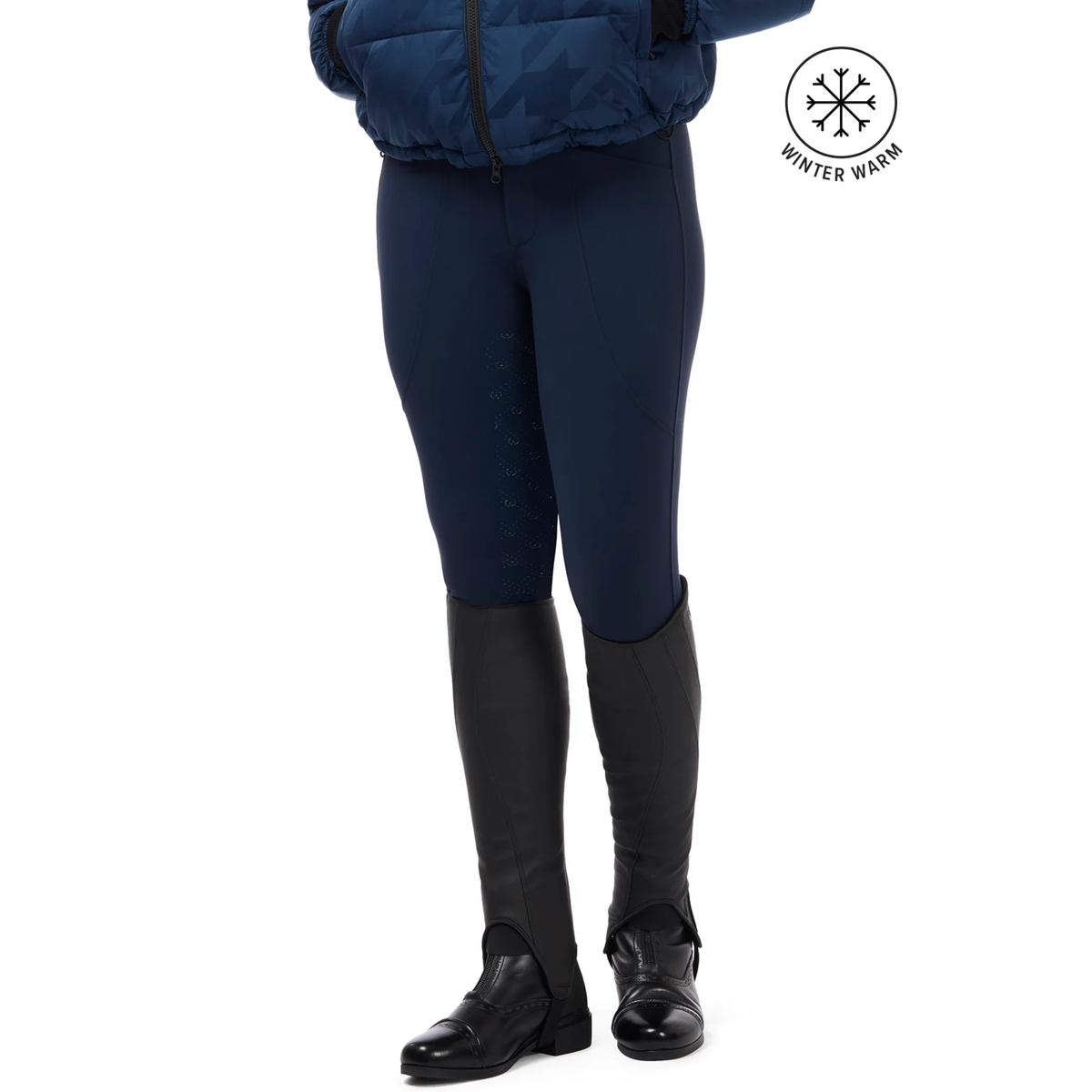 A model from the waist down wearing nightfull colored Kerrits Fusion II Full Leg Equestrian Clini Breeches, black riding boots, and a navy quilted jacket, with a &