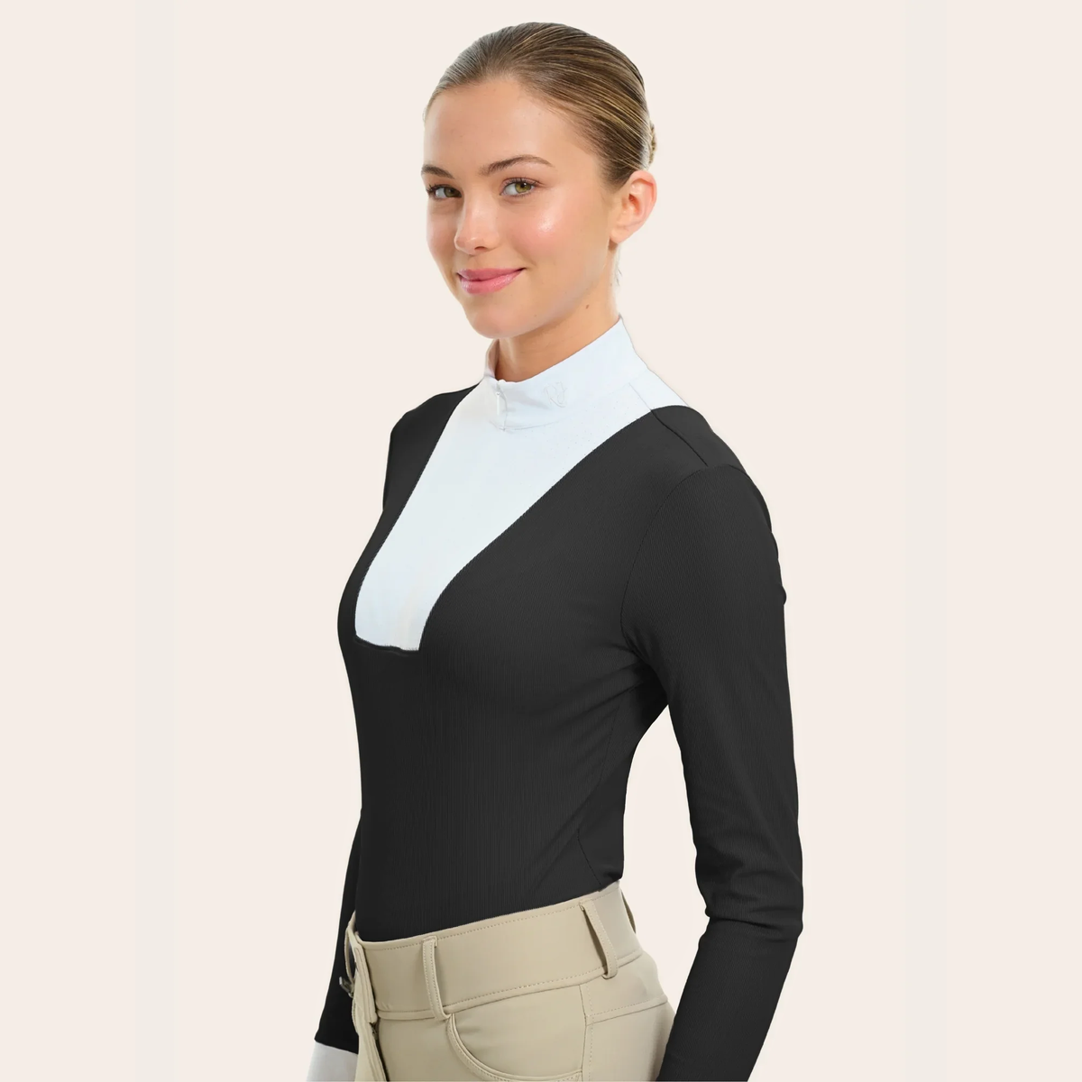 A woman modeling a long-sleeve R.J. Classic Celia Show Shirt with a color-blocked design featuring a black body and a white V-shaped yoke at the neckline, paired with beige riding pants against a light beige background.