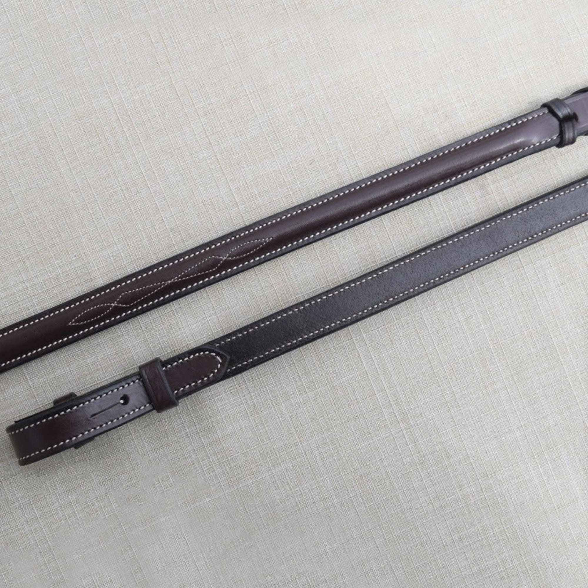 A pair of brown kl select round raised fancy lace reins with white stitching, laid out on a textured background.