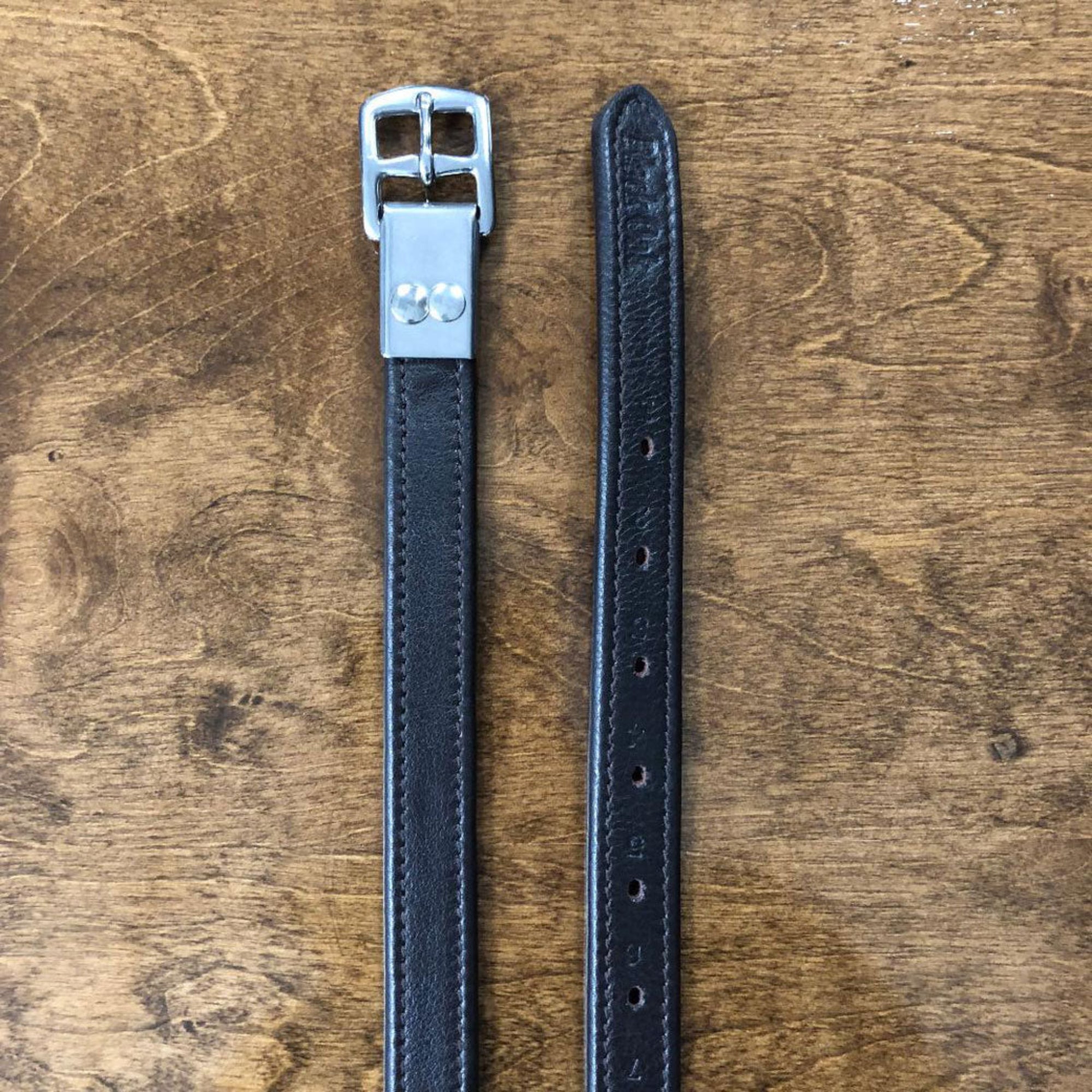 A pair of black kl select black oak riveted calf lined leathers with adjustable holes and a secure riveted buckle, placed on a wooden surface.