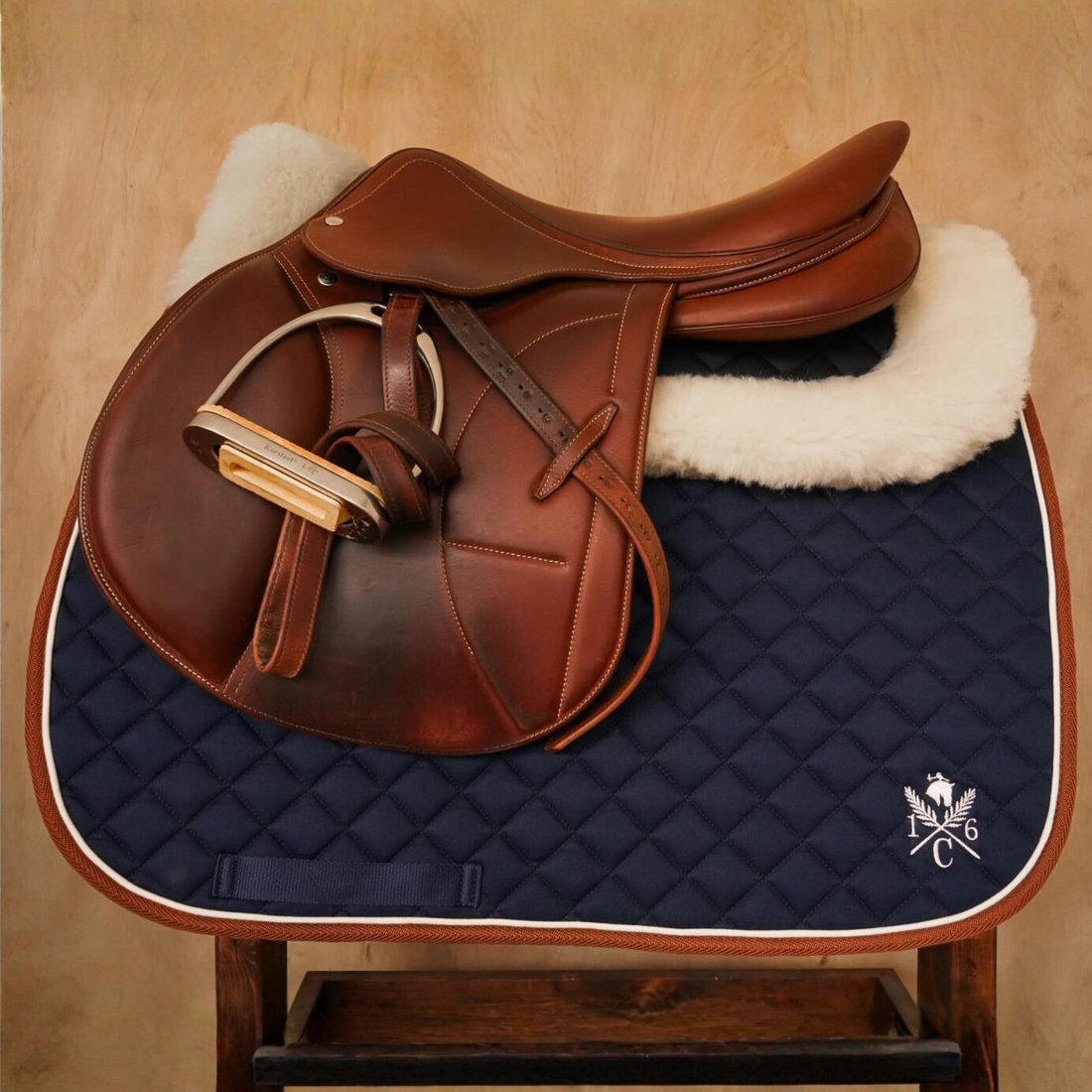 Brown saddle on a navy quilted 16 Cypress Daily Sport Jumper saddle pad with the 16C logo on a wooden stand against a beige paneled background.