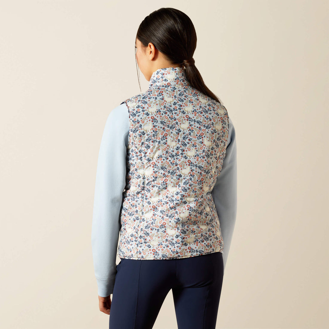 Young lady wearing a floral patterned Ariat Youth Bella Reversible Insulated vest over a light blue shirt and dark pants on a beige background
