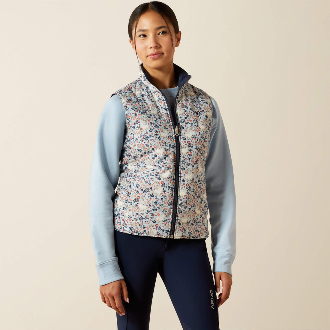 Young lady wearing a floral patterned Ariat Youth Bella Reversible Insulated vest over a light blue long-sleeve shirt and dark pants on a beige background