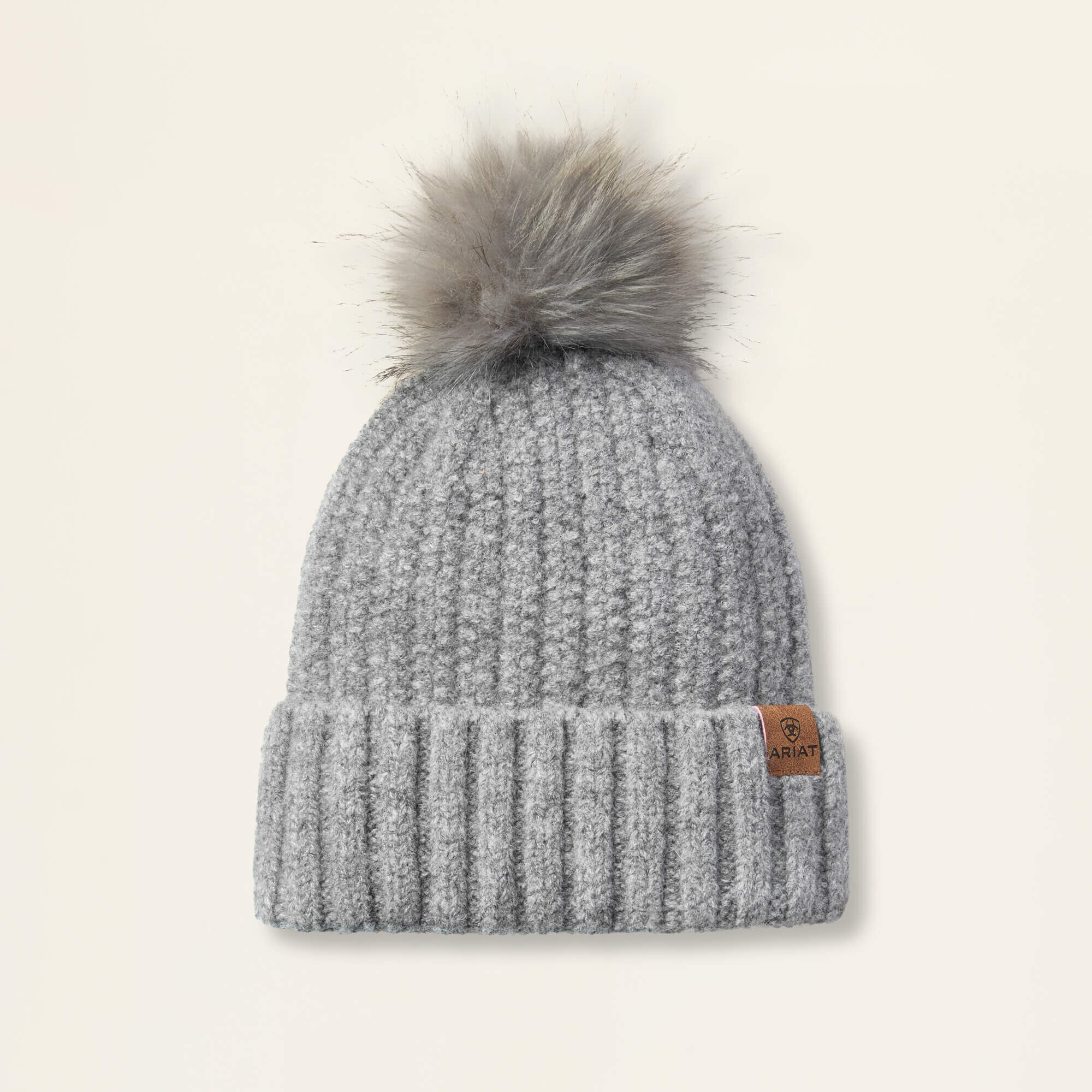 Slate colored Ariat Chiltion knit beanie with a faux fur pom-pom on a beige background