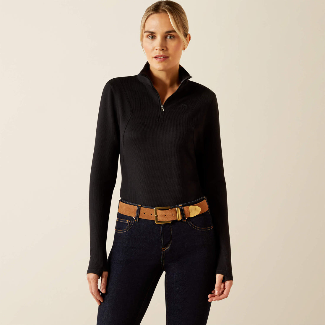 Woman wearing a black long-sleeve. quarter zip Ariat Lucent baselayer and dark jeans with a brown belt on a beige background