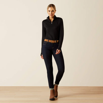 Woman wearing a black long-sleeve, quarter zip Ariat Lucent baselayer, dark jeans, and brown boots on a beige background