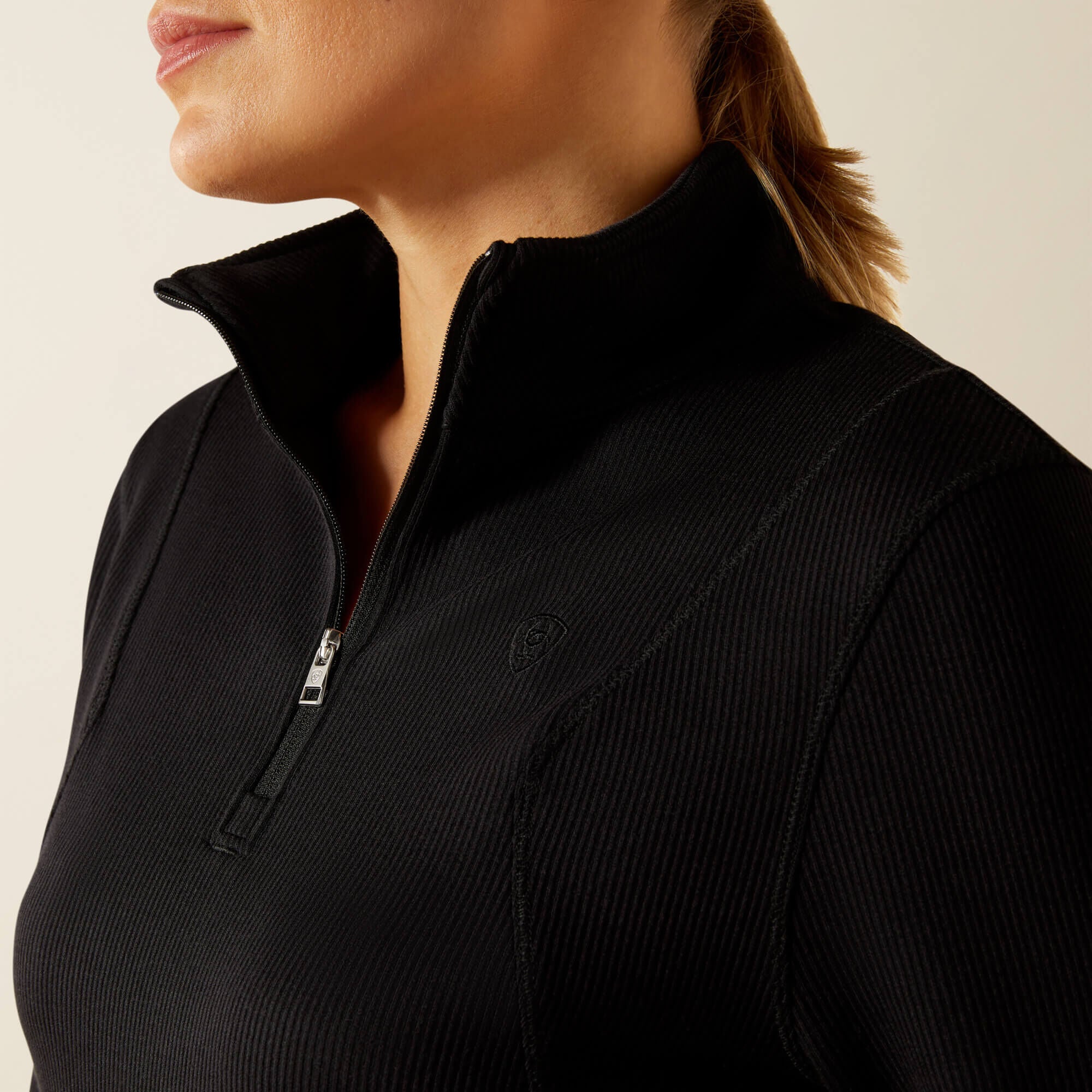Close-up of a woman wearing a black, long sleeve, quarter zip Ariat Lucent baselayer on a beige background