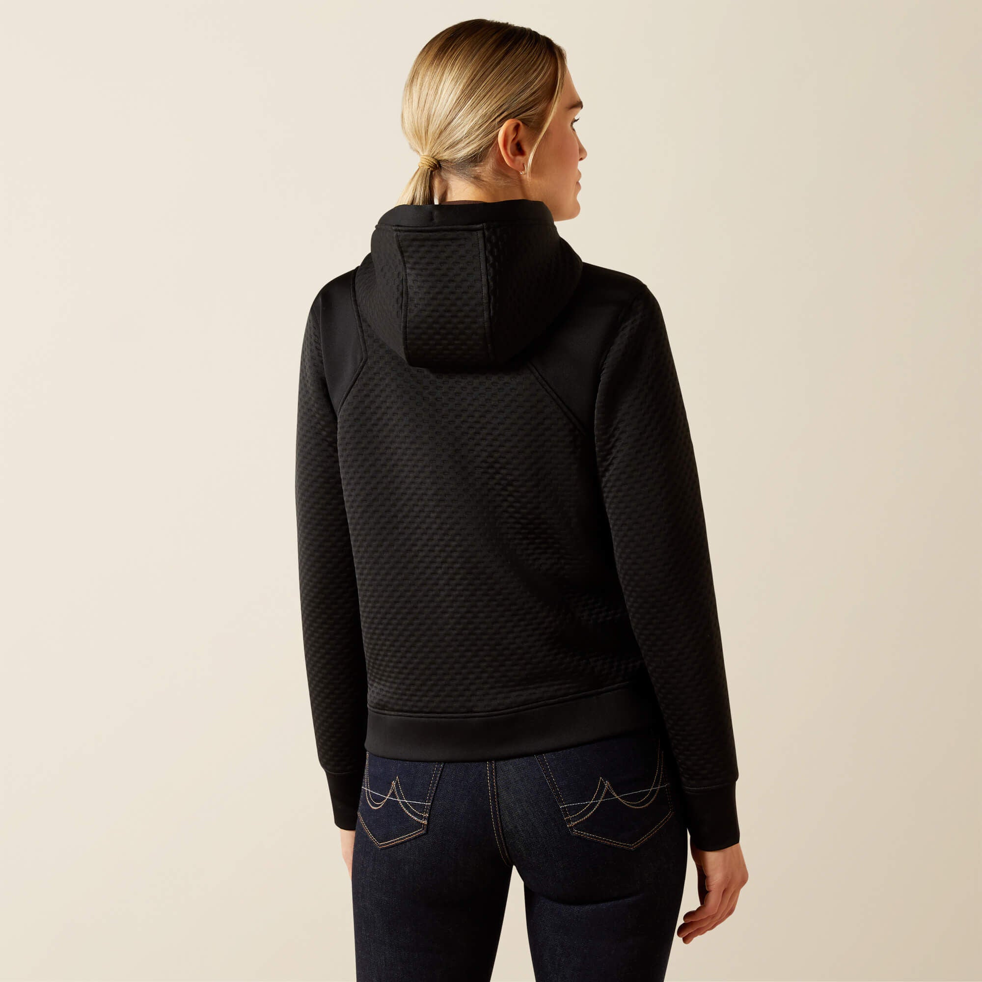 Blonde woman wearing a black patterned Ariat Radiance Full Zip jacke and dark jeans on a beige background