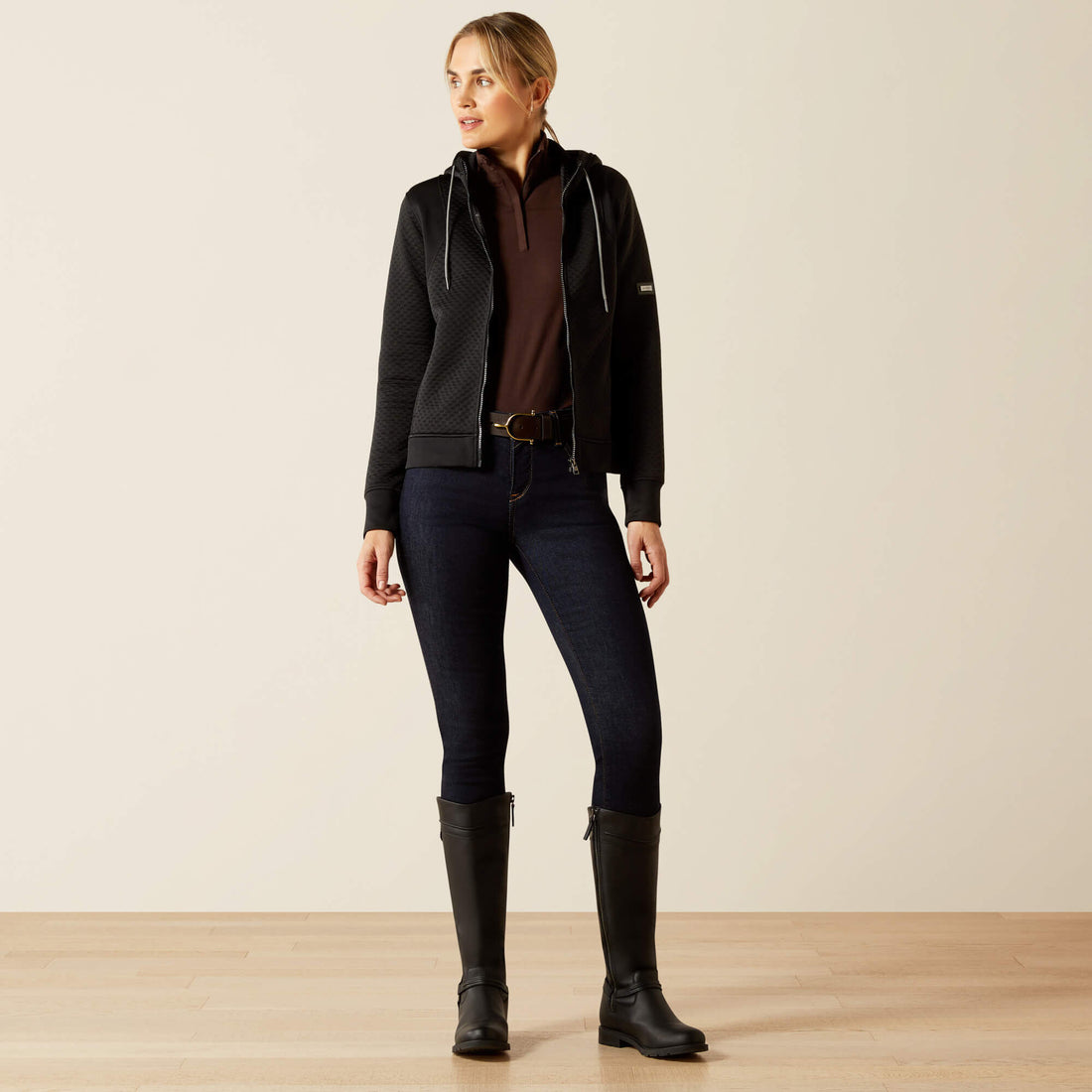 Woman wearing an Ariat Radiance Full Zip black jacket, brown shirt, dark jeans, and black riding boots on a beige background
