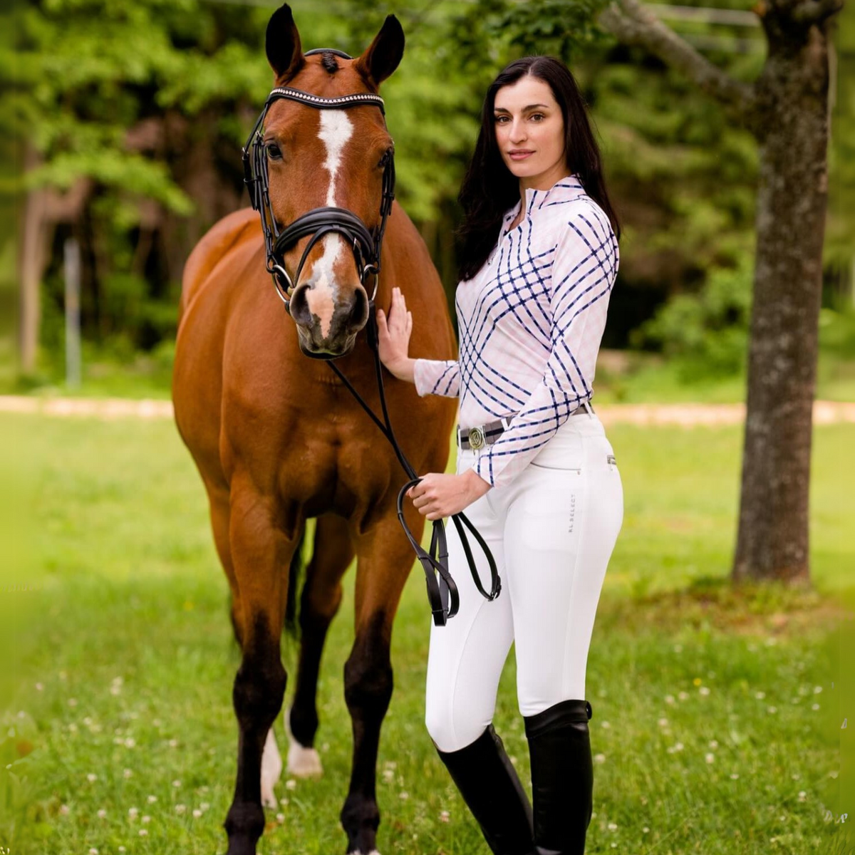 A person wearing white kl gabrielle full seat breeches with black accents, standing next to a brown horse, in a grassy field.