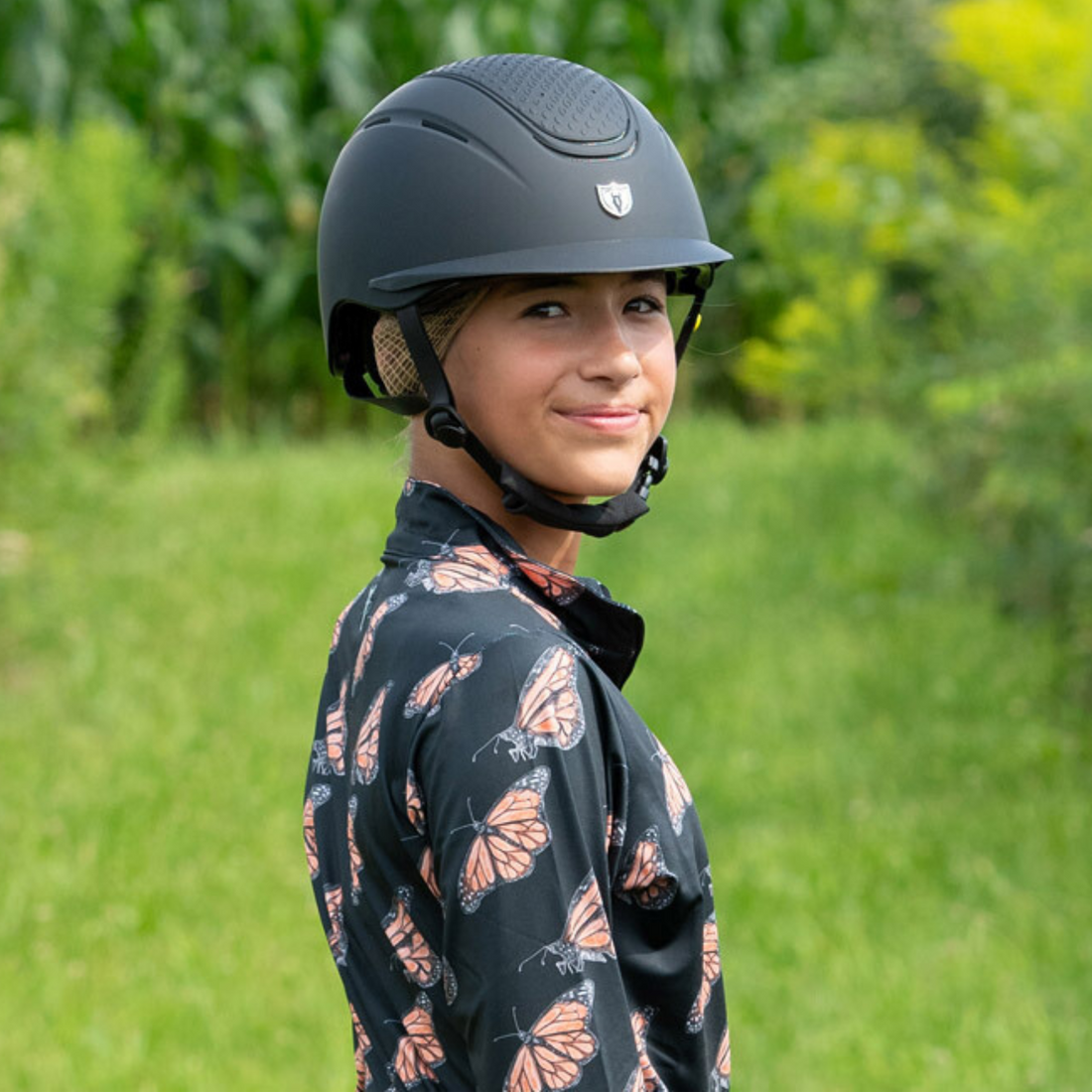 Lifestyle image of a female equestrian wearing Tipperary black matte Clover Mips Helmet while standing in a grassy field. 