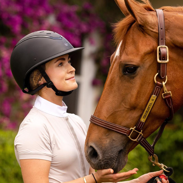 Lady wearing a EQx by Charles Owen Kylo Black Matte with Black Gloss Frame  Riding Helmet.  She is holding a chestnut horse wearting a brown leather halter.