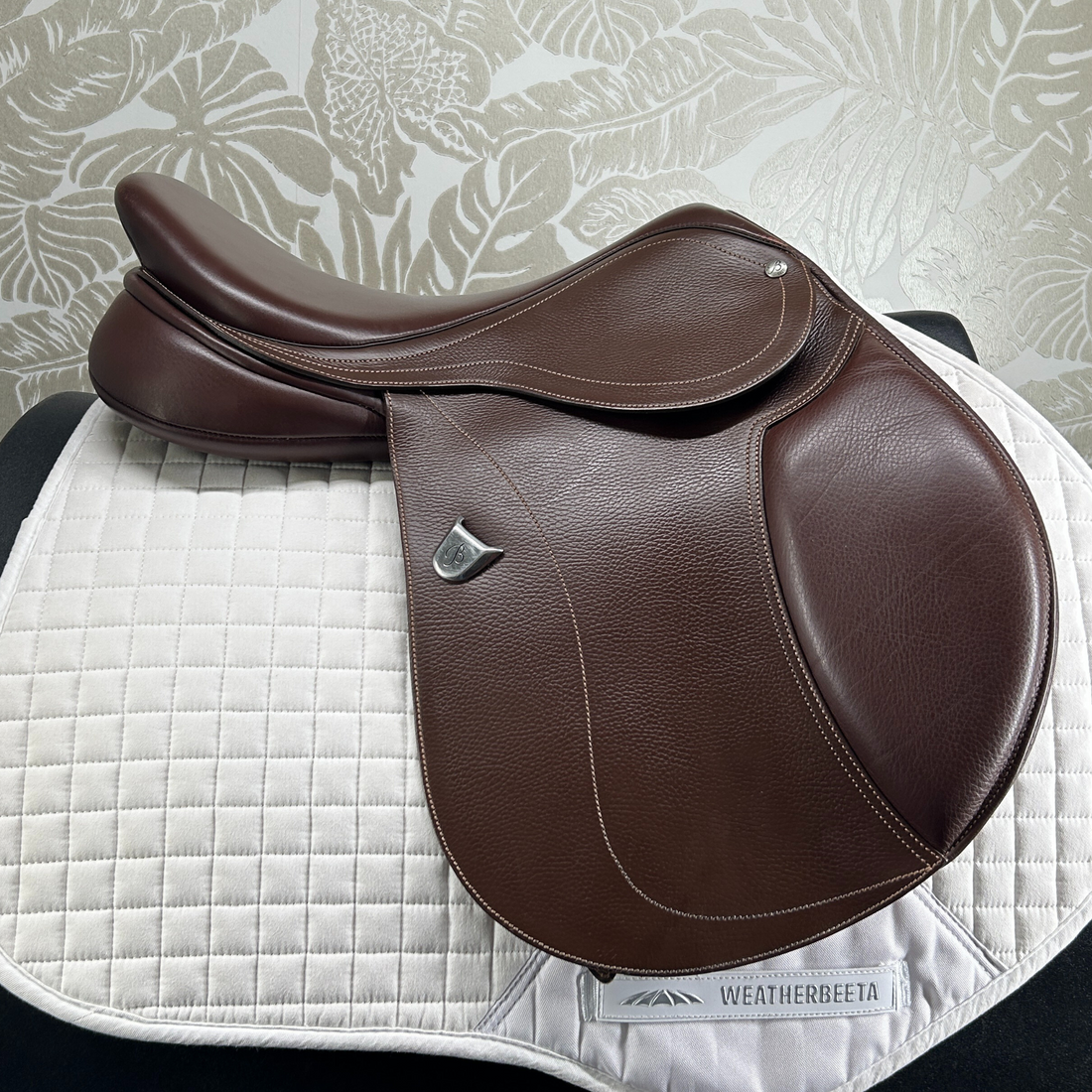 Brown used 17&quot; Bates Hunter Jumper saddle on a white quilted pad with against a floral-patterned wall.