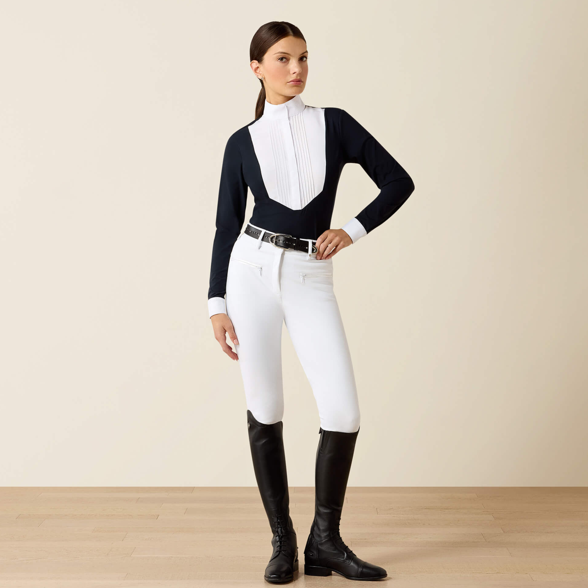 Woman wearing a navy Ariat SolVeil Long Sleeve Show Shirt with a white bib front  and white breeches and tall black field boots on a beige background .