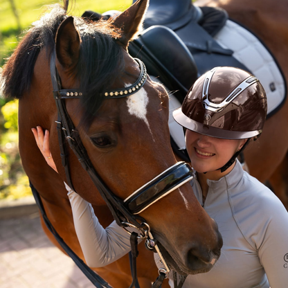 A smiling equestrian in BR Equestrians Zeta Serene Shine brown riding helmet and light gray riding shirt stands beside a brown horse wearing a black bridle. The rider is touching the horse&