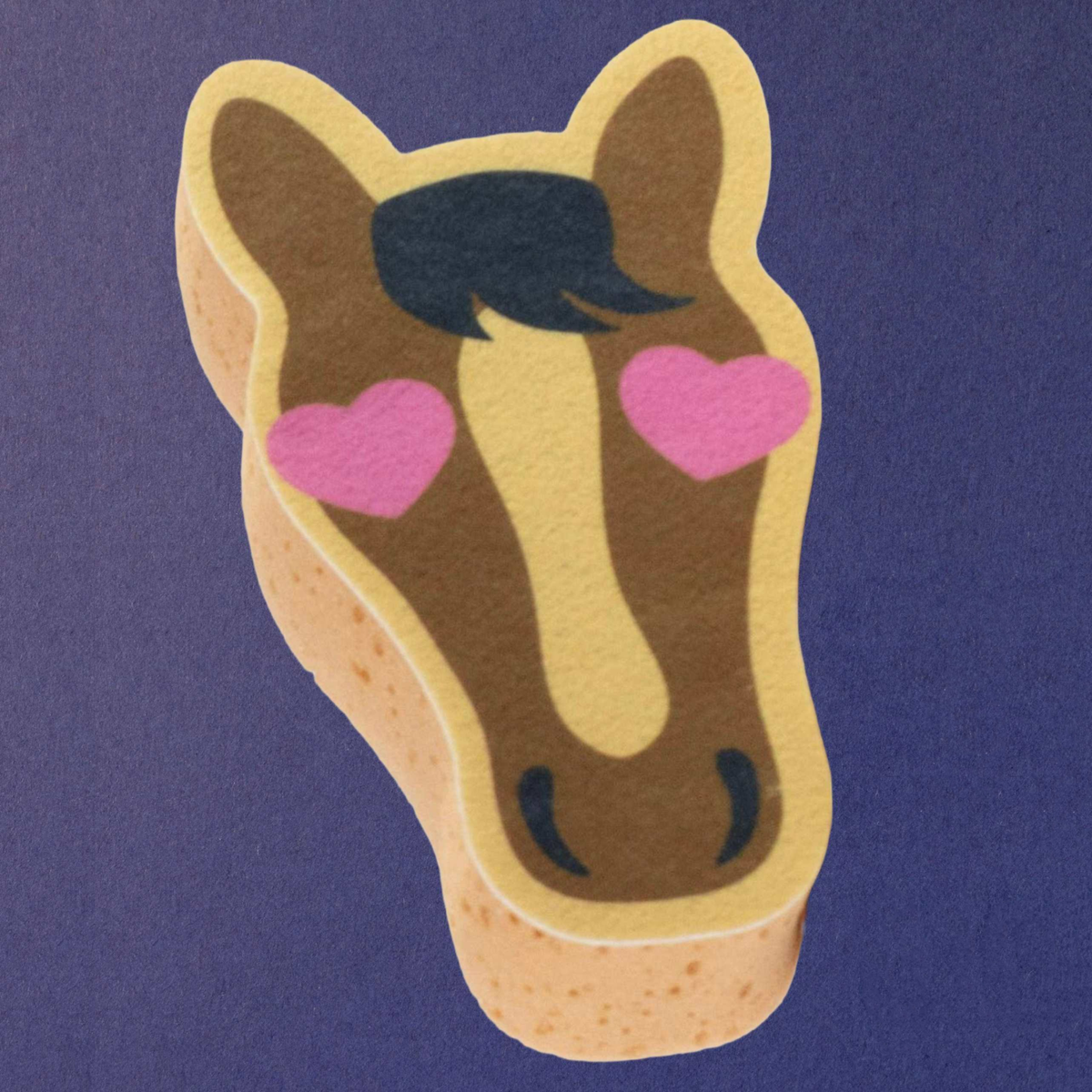 Dreamers and Schemers Pony Love novely horse bath spong brown face, beige outline, black mane, and pink heart-shaped eyes, shown on a solid purple background.