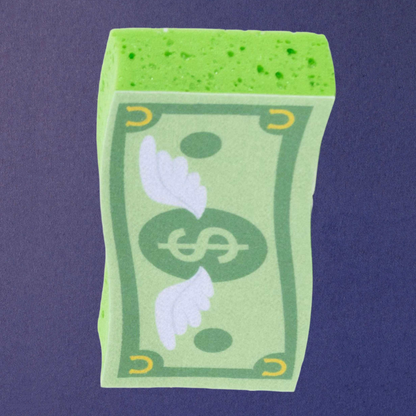 Dreamers &amp; Schemers novelty rectangular cash money sponge shaped like a stylized green dollar bill, featuring a white winged dollar sign in the center, yellow corner horseshoe accents, and a bright green porous top surface, shown against a solid dark purple background.