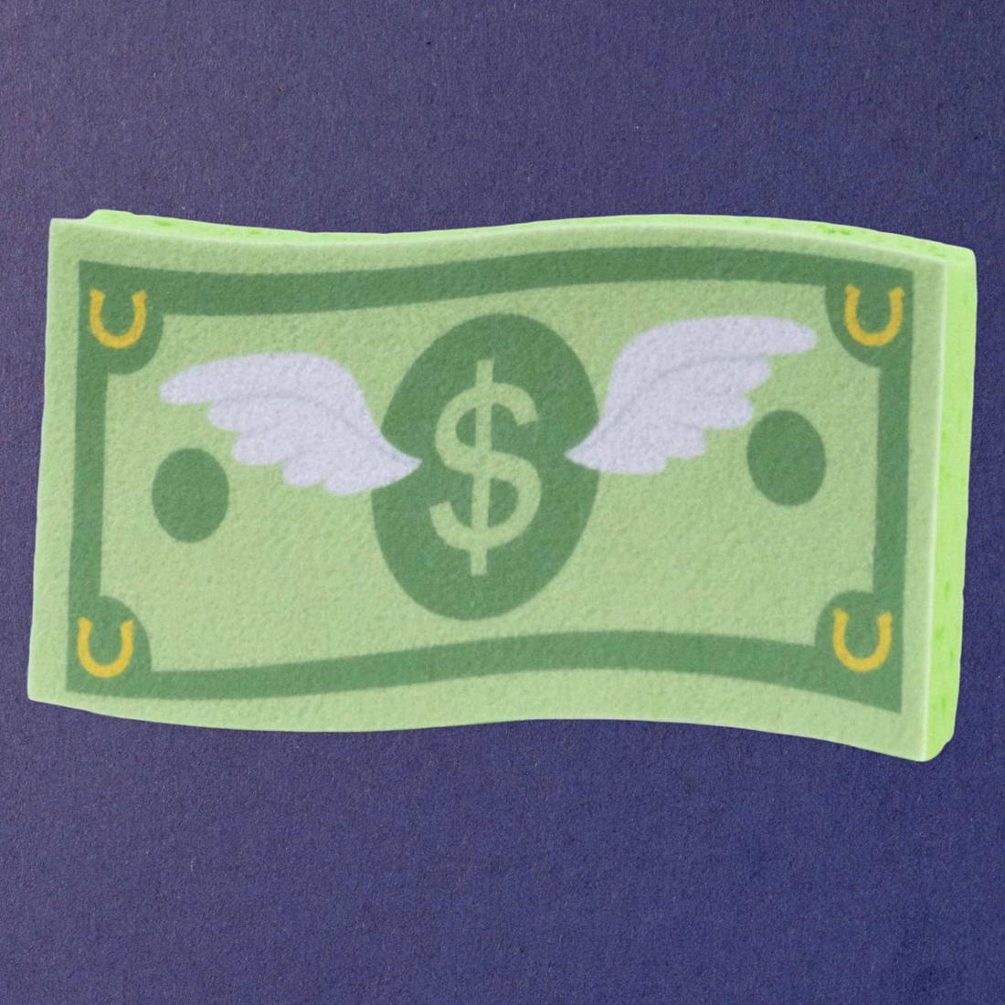 A green dreamers &amp; schemers money bill shaped bath sponge with white wings and a dollar sign in the center, displayed on a dark purple textured background.