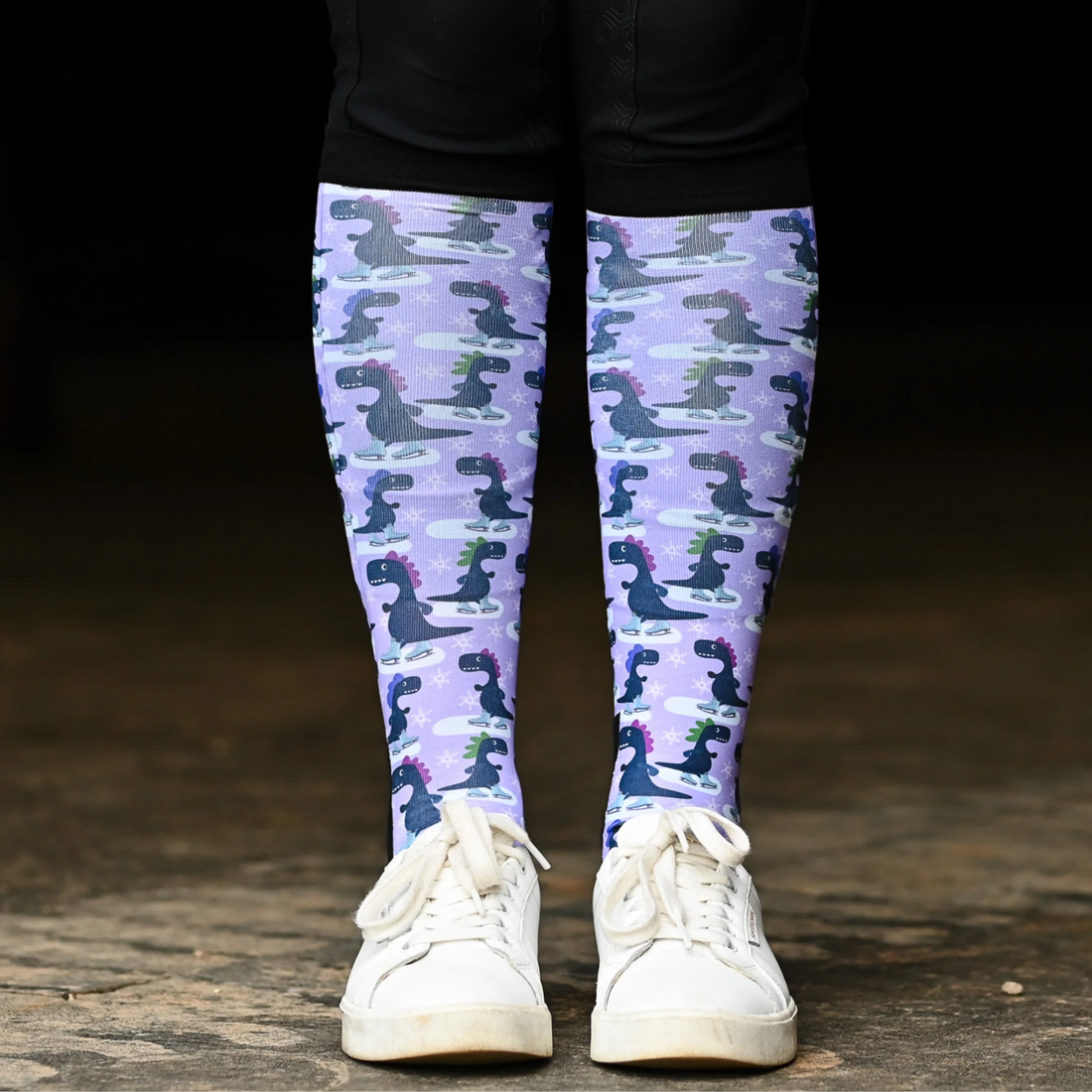 A rider wearing Dreamers &amp; Schemers Dino-Skate boot socks with a purple and gray dinosaur ice skating pattern, paired with white sneakers and black breeches.