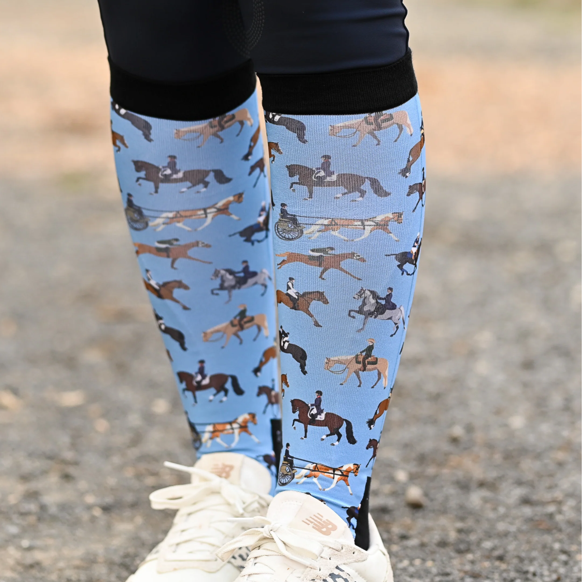 A person outdoors wearing Dreamers &amp; Schemers The Pony Island "Horse Sports" tall boot socks with an all-over equestrian print of horses and riders, paired with black breeches and white sneakers.