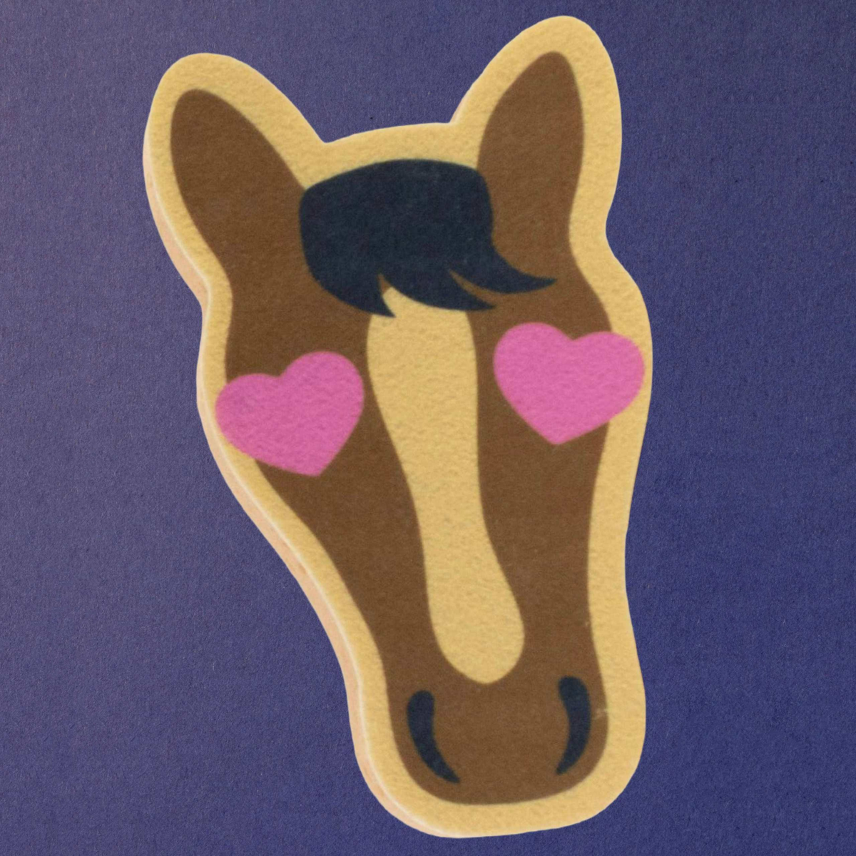 A novelty equine sponge shaped like a horse head, with a brown body, beige outline, black mane, and pink hearts for eyes, shown on a textured purple background.