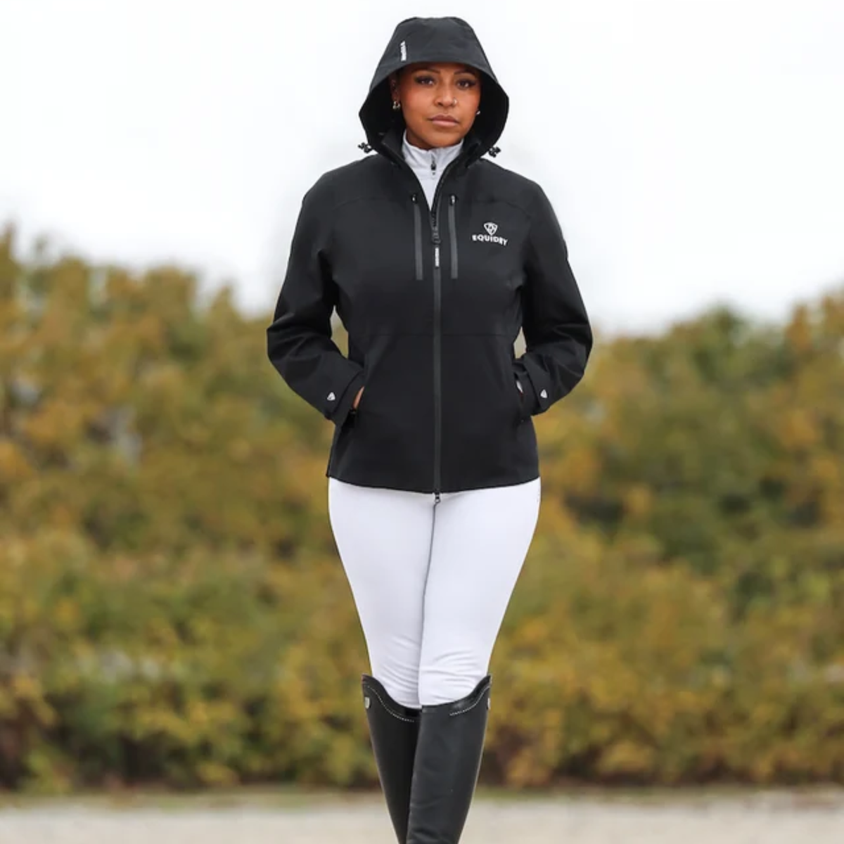 A woman wearing a black hooded EQUIDRY Varsha coat with a white logo on the left chest, paired with white riding breeches and tall black riding boots, standing outdoors against a blurred background of green and yellow trees.