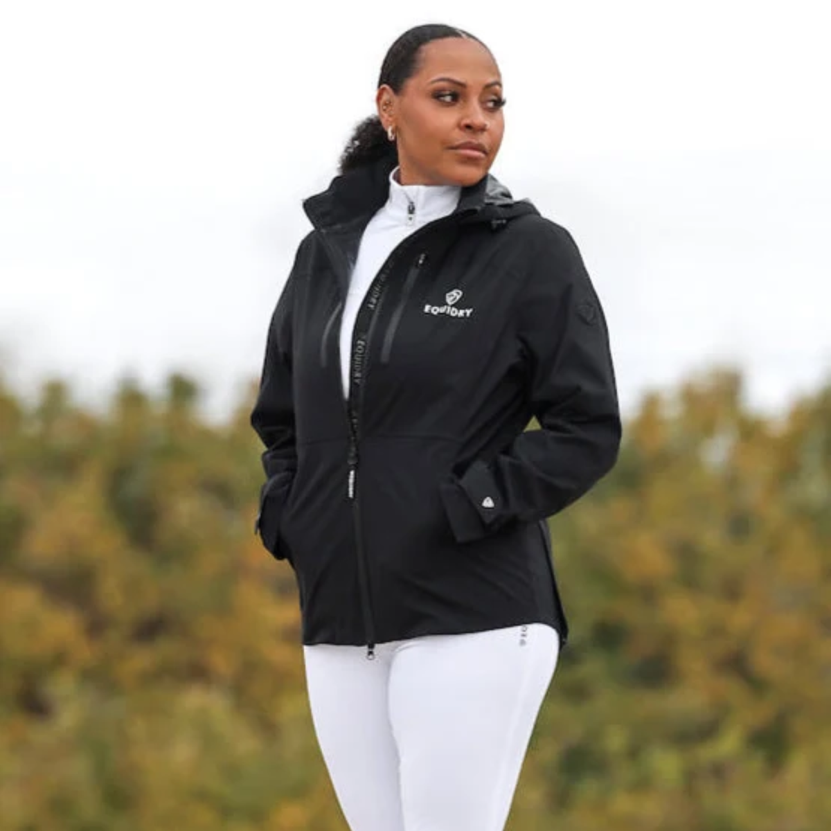 A woman wearing a black EQUIDRY Varsha coat with a white logo on the left chest, layered over a white top and white breeches, standing outdoors against a blurred background of trees.