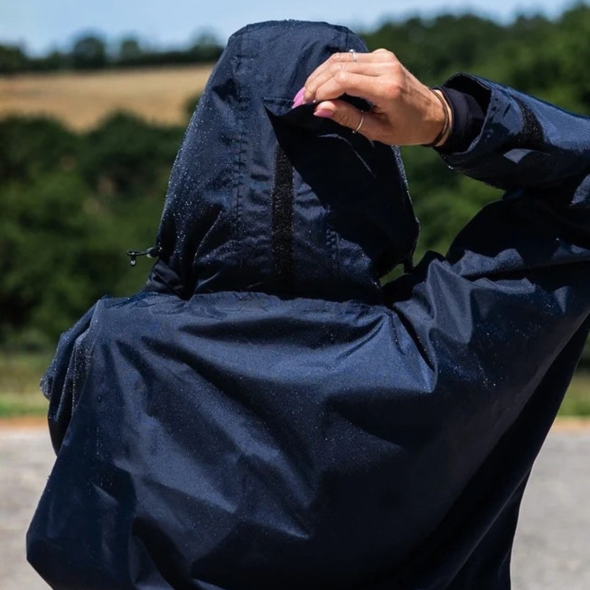 A person wearing a EquiDry Evolution Mesh Lined navy hooded coat, viewed from behind, with their hand adjusting the hood. The background shows a blurred outdoor landscape with green trees and a field.