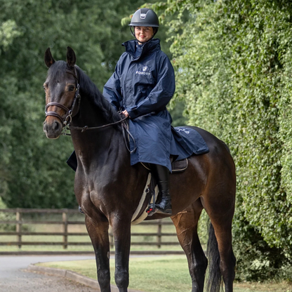 A rider wearing a navy blue EquiDry Evolution Mesh Lined Coatt and helmet rides a dark brown horse outdoors on a path with green trees in the background. The raincoat has a white "EquiDry" logo on the chest.