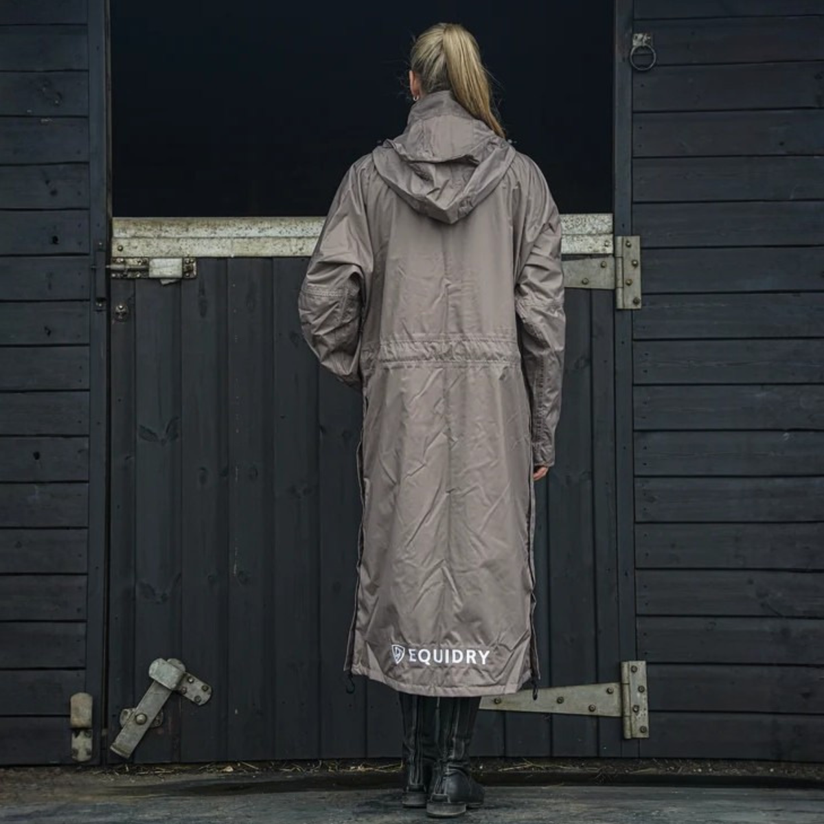 A woman stands with her back to the camera wearing a long taupe EquiDry Evolution Mesh Lined Coat  in front of a dark wooden stall door. The brand name &