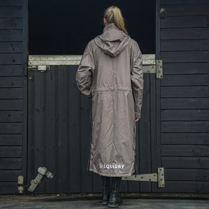 A woman stands with her back to the camera wearing a long taupe EquiDry Evolution Mesh Lined Coat  in front of a dark wooden stall door. The brand name &