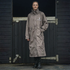 A woman stands in front of a wooden stall door wearing a long taupe EquiDry Evolution Mesh Lined waterproof coat with a white &