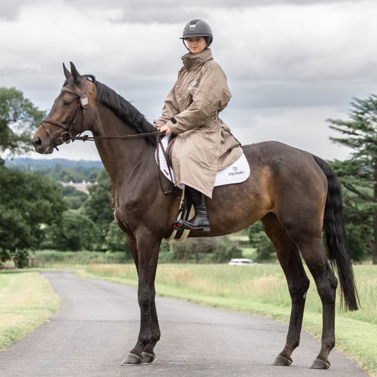 A woman riding a dark brown horse on a british road, wearing a taupe EquiDry Evoulution Mesh Lined coat and black riding helmet. The horse has a white saddle pad with the brand name &