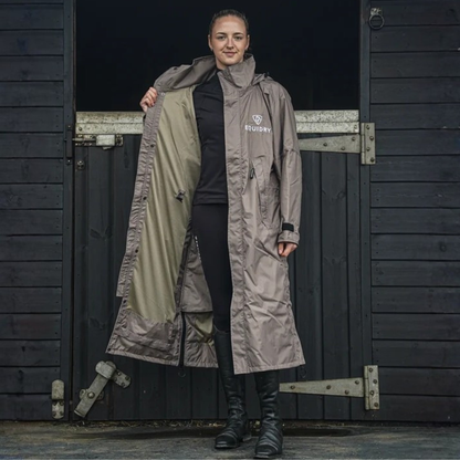 A woman stands in front of a wooden stall door wearing a long EquiDry Evolution Mesh Lined waterproof coat. The coat is a taupe with a light olive interior lining, and features the &