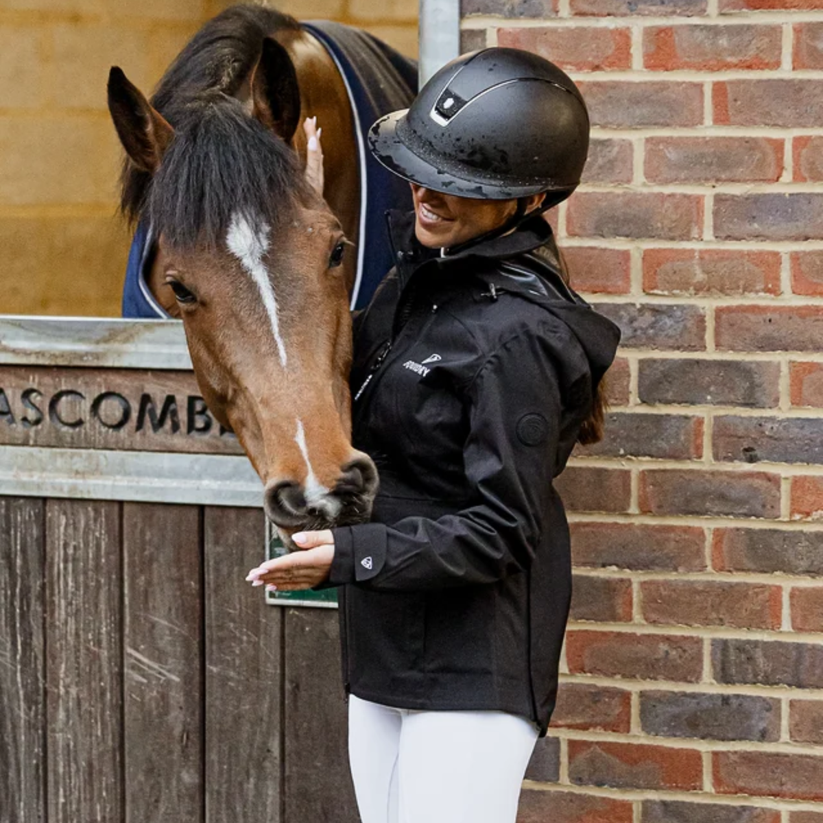 A woman in equestrian attire, wearing a black riding helmet and a black EQUIDRY Varsha coat stands next to a brown horse in a stable while feeding it.
