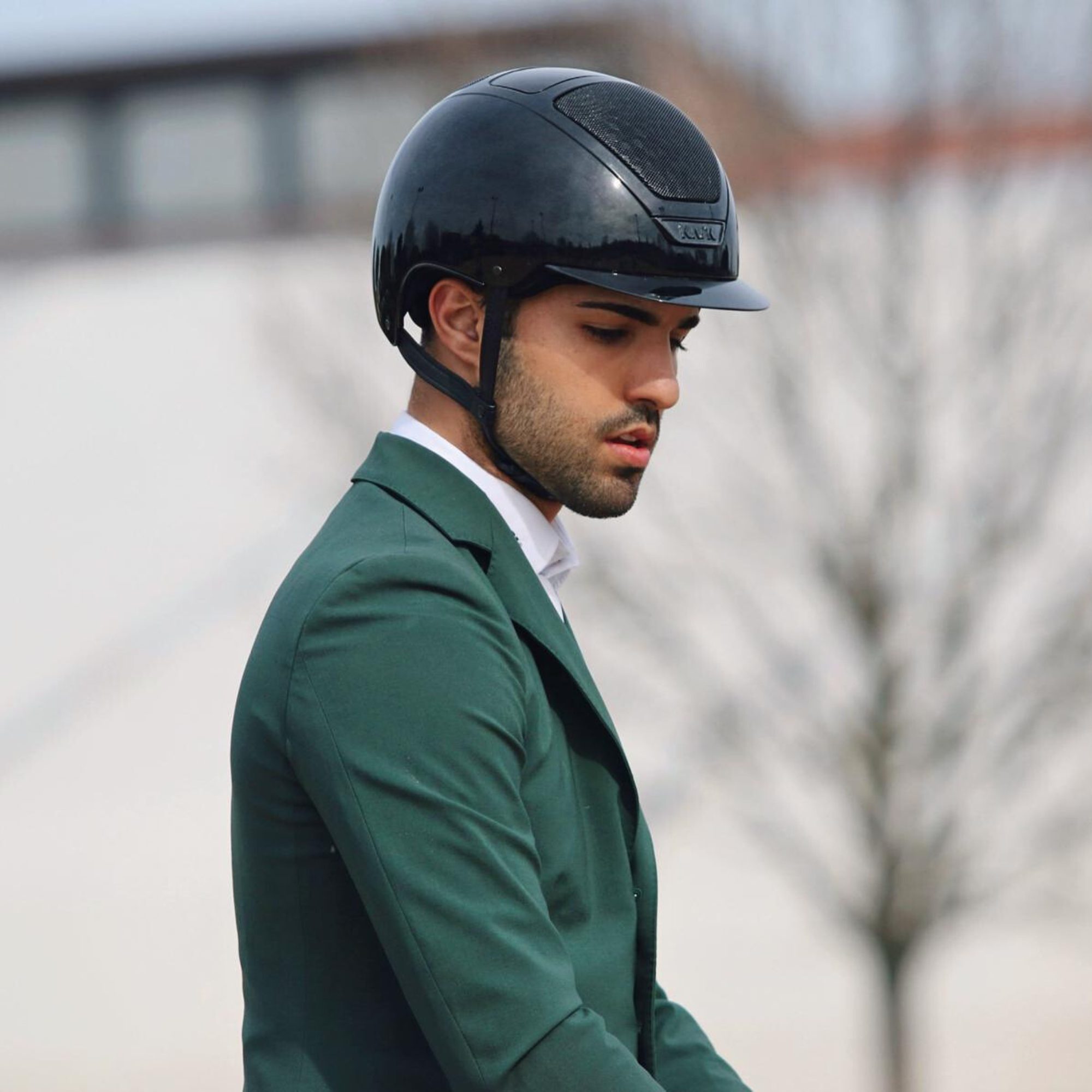 Male rider wearing a KASK Dogma Pure Shine black riding  helmet and green equestrian show coat outdoors.