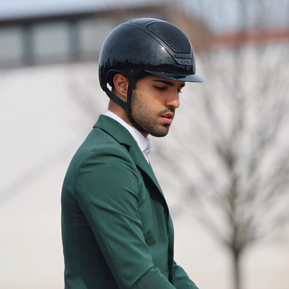 Male rider wearing a KASK Dogma Pure Shine black riding  helmet and green equestrian show coat outdoors.