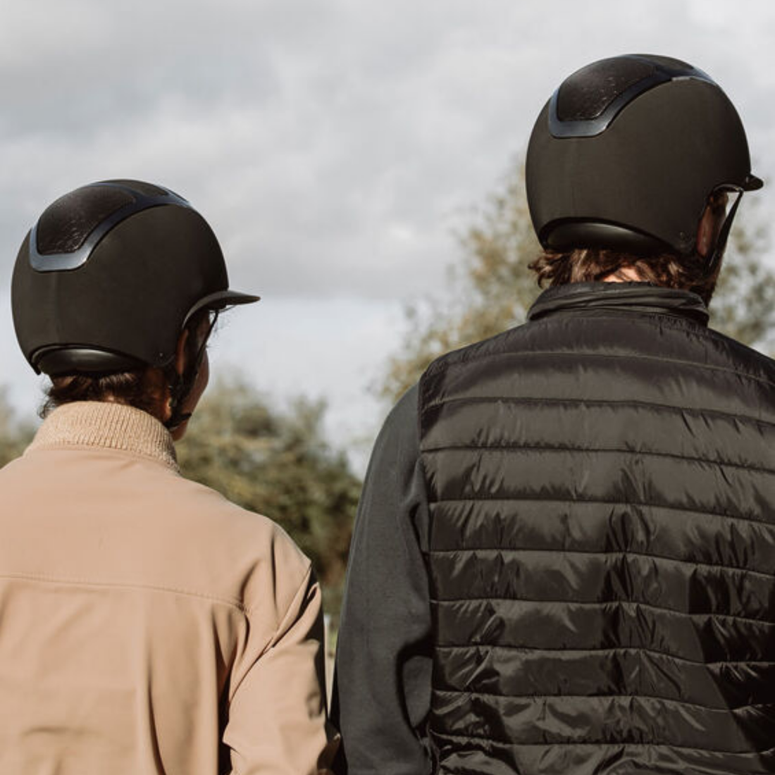 Two riders seen from behind wearing matte black KASK riding helmets with KASKs comfort fit system,  and outerwear, standing outdoors under an overcast sky.